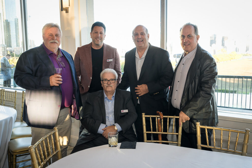 Five men posing for group photo at corporate event with city skyline visible through windows