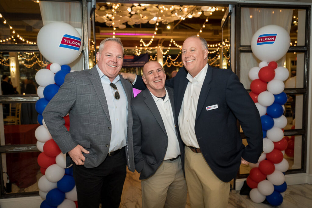 Three men smiling together under Tilcon-branded balloon archway at corporate event