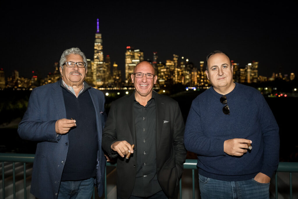 Three executives with cigars on outdoor terrace with Manhattan skyline glowing behind them
