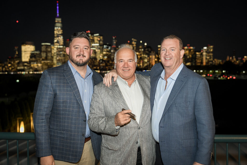 Three men posing with cigars on terrace with illuminated NYC skyline at night