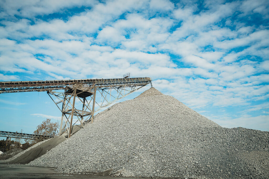 Conveyor belt system loading large conical aggregate pile under partly cloudy sky at Clinton Point Quarry.
