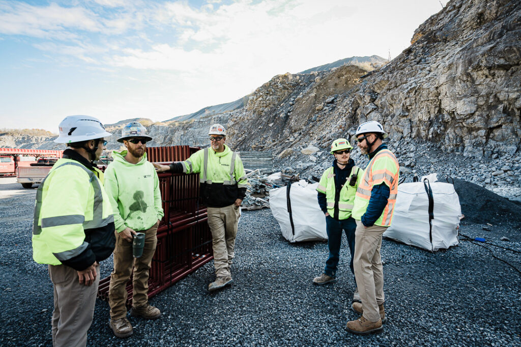 Tilcon team members in safety gear having discussion in quarry pit with material piles and hillside behind them