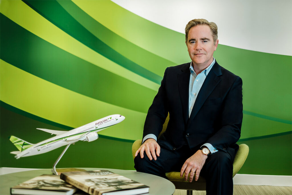 Corporate executive portrait of a man in a suit seated next to a model airplane in a modern office environment.