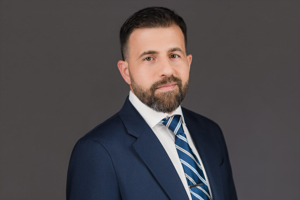 Professional corporate headshot of a man in a navy suit and striped tie against a neutral gray background, photographed by Alex Kaplan Photo in New Jersey.