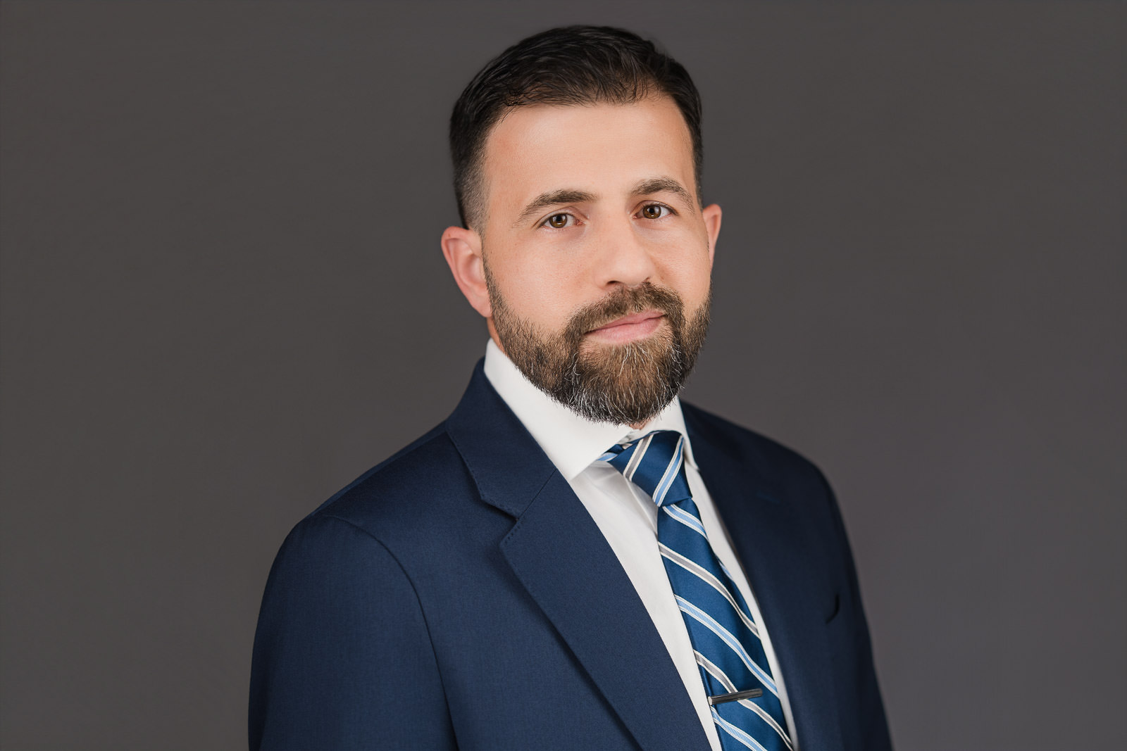 Professional corporate headshot of a man in a navy suit and striped tie against a neutral gray background, photographed by Alex Kaplan Photo in New Jersey.