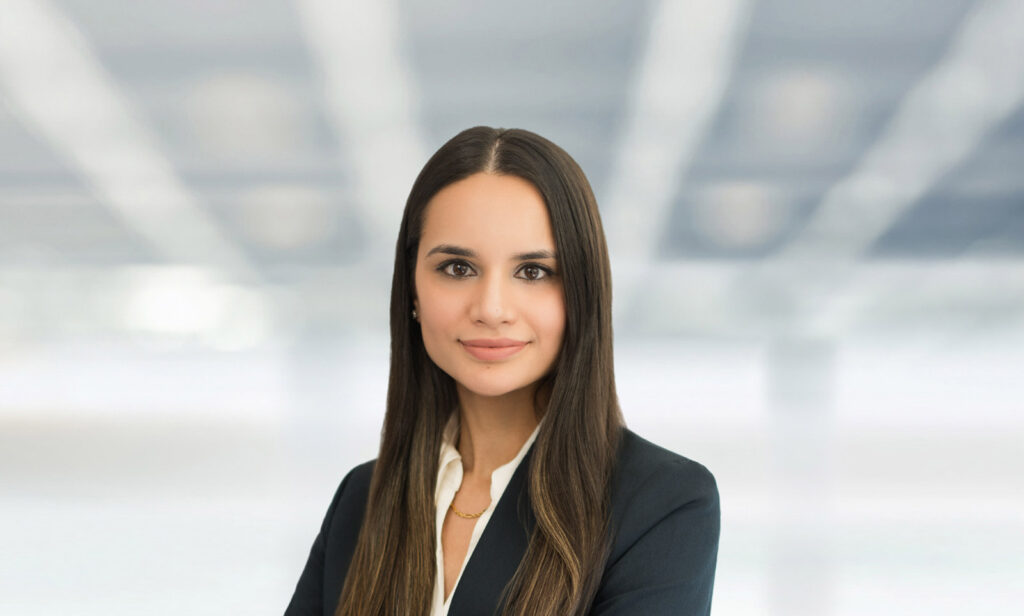 Professional corporate headshot of a young woman in a dark blazer photographed against a blurred architectural ceiling background, taken by Alex Kaplan Photo in Northern New Jersey.