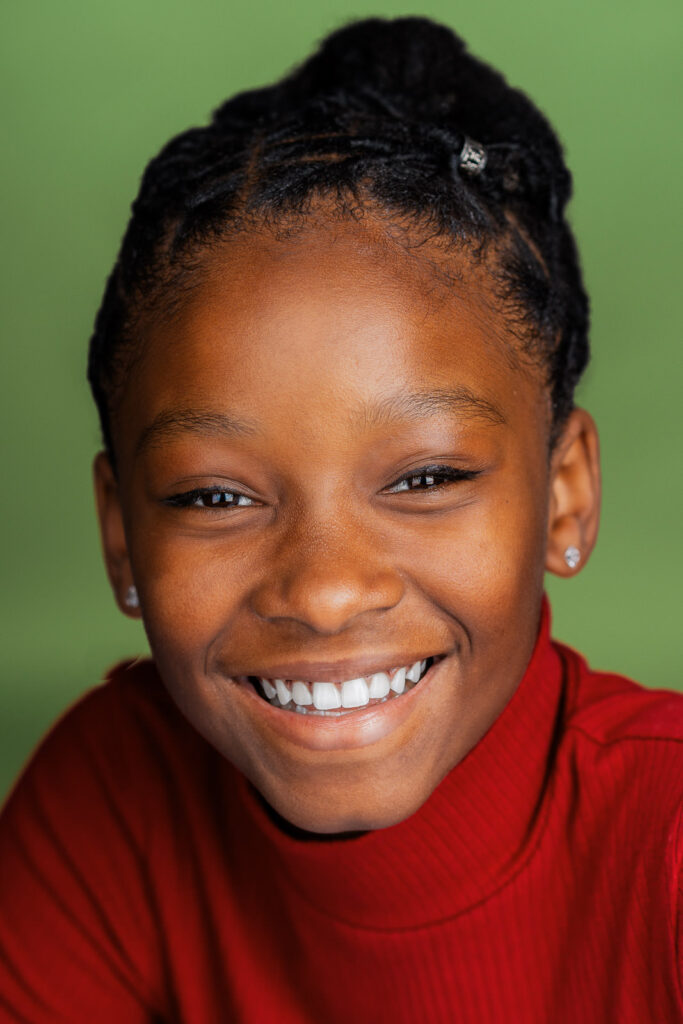 Young girl with braided bun looking over shoulder at camera during professional actor headshot session in New Jersey studio, wearing denim jacket against yellow background.
