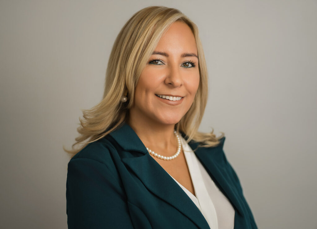 Professional corporate headshot of a woman in a teal blazer and pearl necklace smiling against a neutral gray background, photographed by Alex Kaplan Photo in Northern New Jersey.