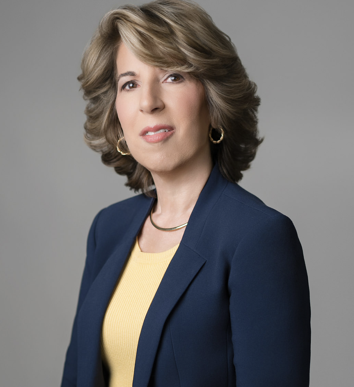 Professional corporate headshot of a woman wearing a navy blazer and yellow blouse in a studio