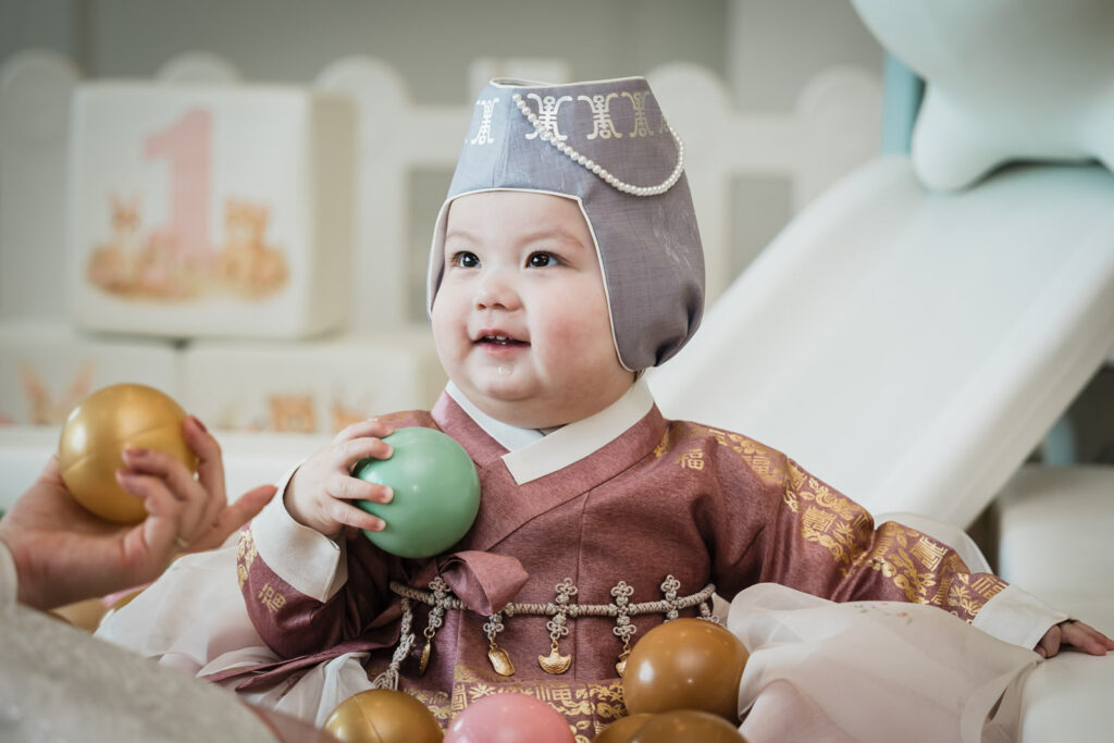 Baby in traditional Korean hanbok holding a ball and smiling at a first birthday celebration in Northern New Jersey