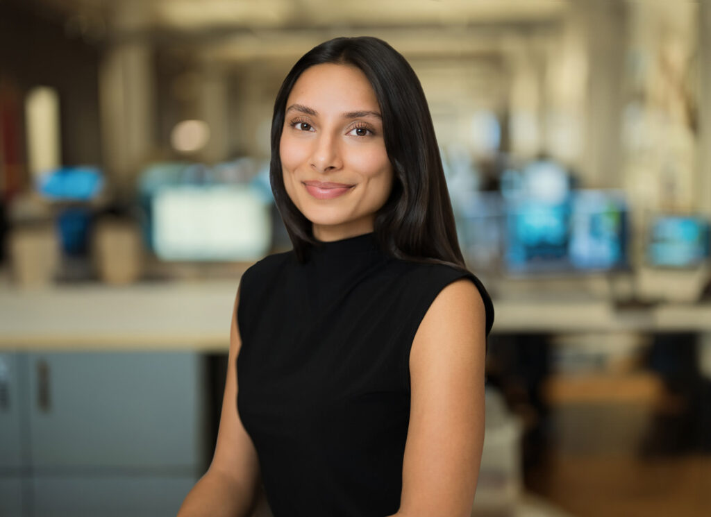 Business headshot of a woman with long dark hair in a black sleeveless turtleneck photographed at a New York City company office Caption (optional): Business headshots NYC — uniform lighting and framing across the full team