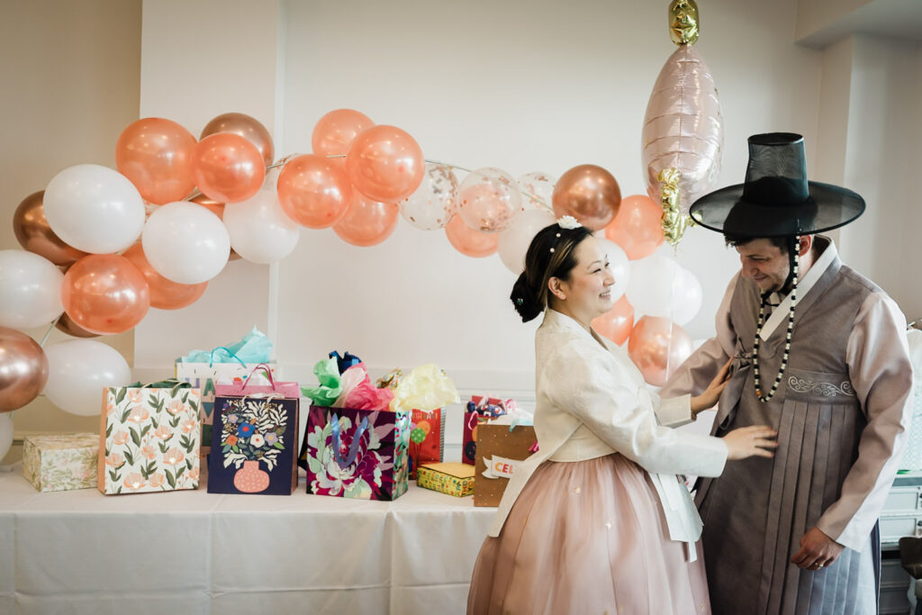 Parents in traditional Korean hanbok sharing a candid moment near the gift table at a first birthday party in New Jersey