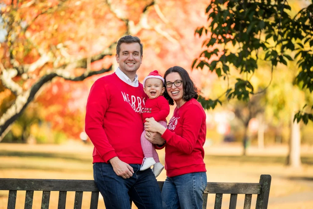 Family of three in matching red Christmas sweaters laughing together at Taylor Park in Millburn New Jersey during fall foliage season