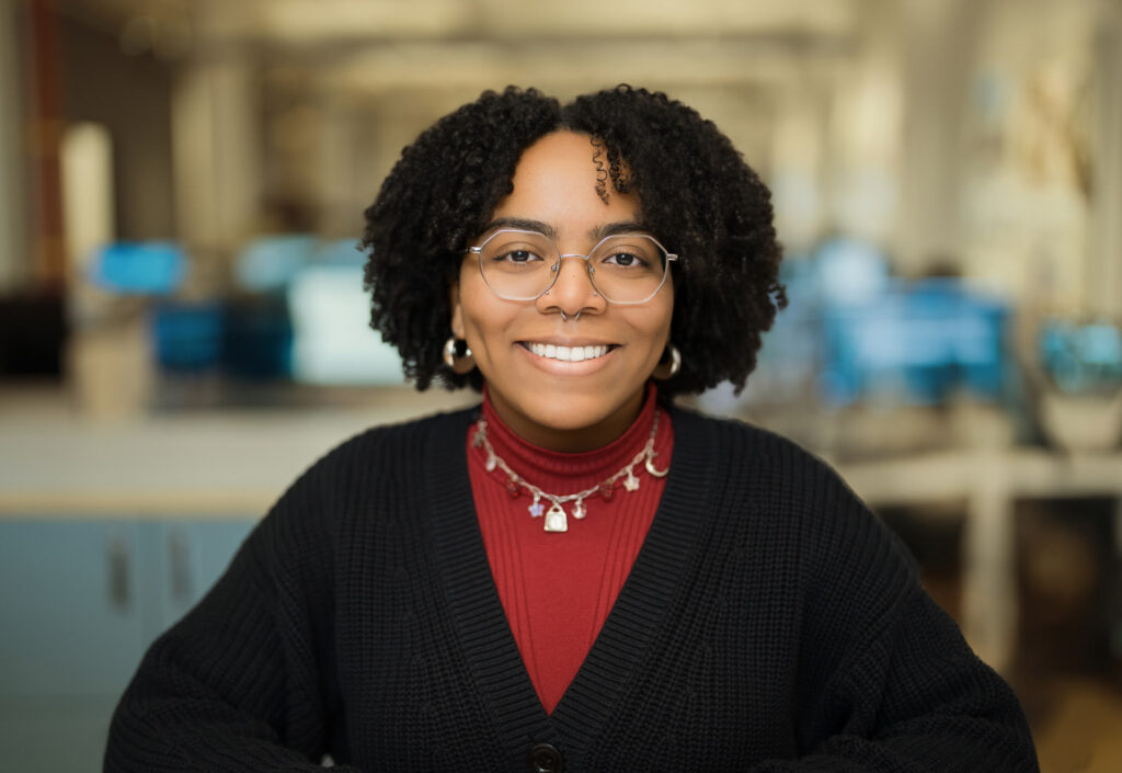 Company headshot of a woman with natural hair and glasses wearing a red turtleneck and black cardigan in a NYC office setting Caption (optional): Company headshots NYC — six team members, one consistent look
