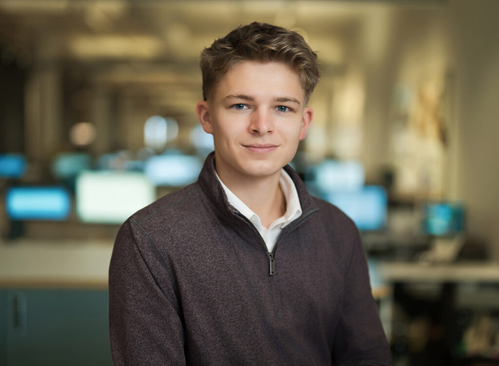Consistent corporate headshot of a young professional in a maroon quarter-zip photographed in a NYC office with soft background blur Caption (optional): Same setup, every person — corporate team headshots NYC