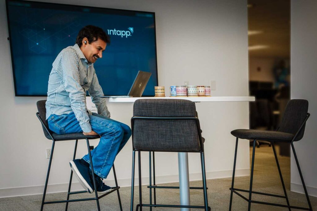 A tech executive laughs while working at a standing table in a modern office, with a branded company screen visible in the background, photographed by New Jersey corporate photographer Alex Kaplan.