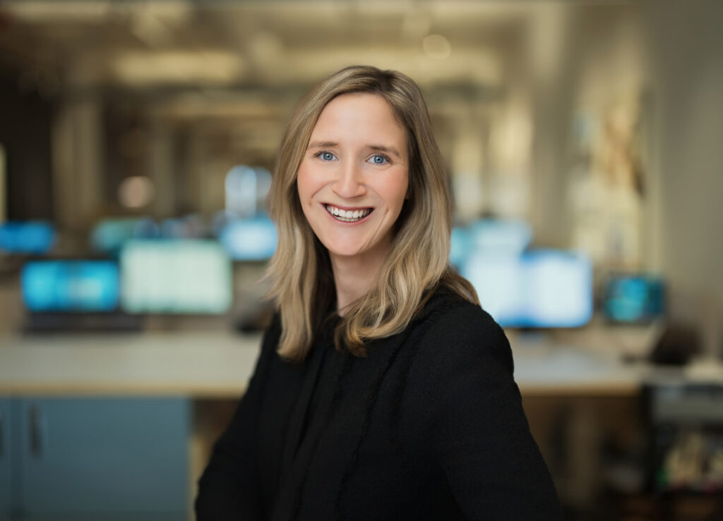 Corporate headshot of a woman in a black blazer photographed in a NYC technology office with blurred monitors in background Caption (optional): Corporate headshots NYC — on-location team session