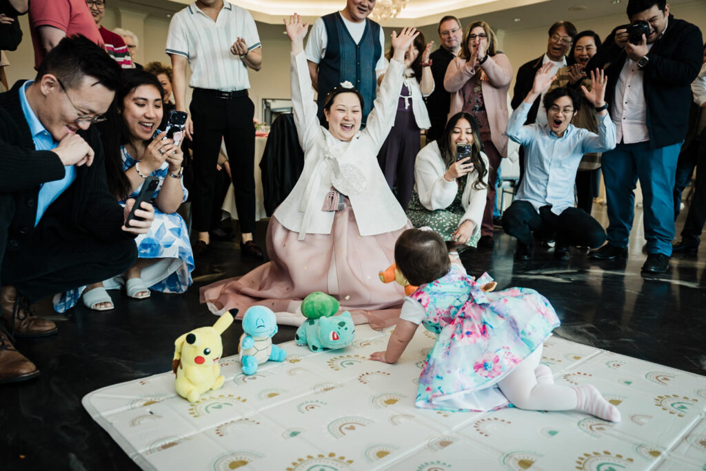 Baby crawling toward Doljabi objects as mother in Korean hanbok reacts with the crowd at a first birthday celebration in New Jersey