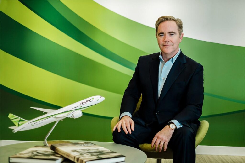 Corporate executive portrait of a man in a suit seated next to a model airplane in a modern office environment.
