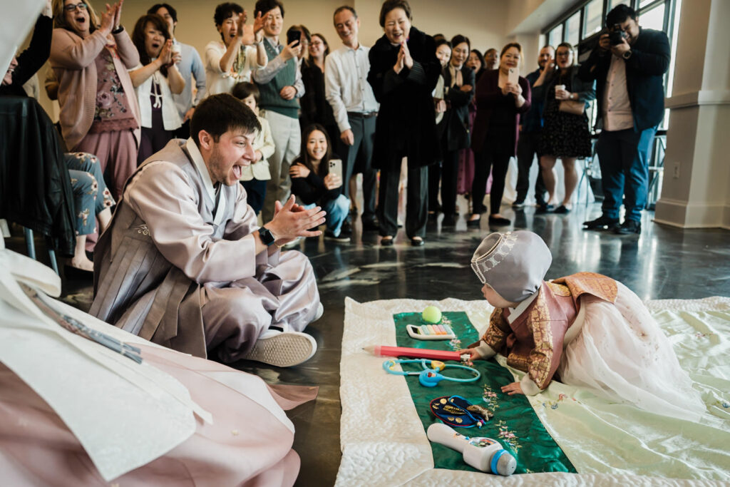Father cheering with excitement as baby reaches toward traditional Doljabi objects at a Korean first birthday party in New Jersey