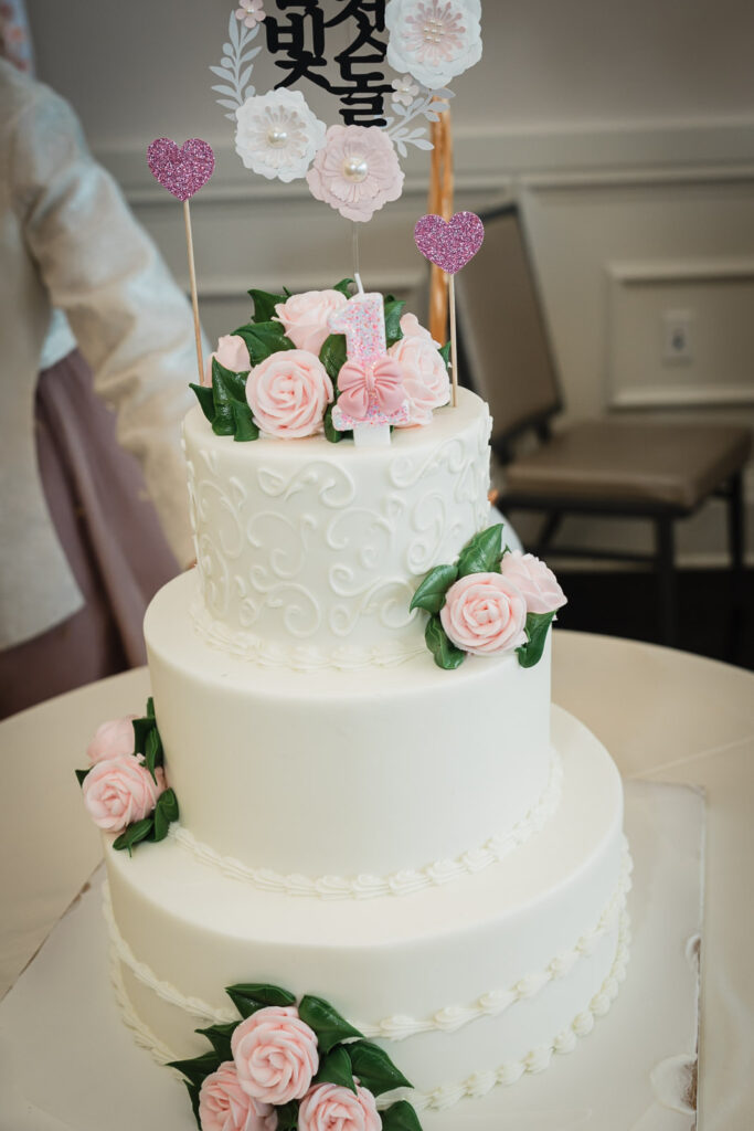Three-tier white first birthday cake with pink roses and Korean birthday topper at a celebration in New Jersey