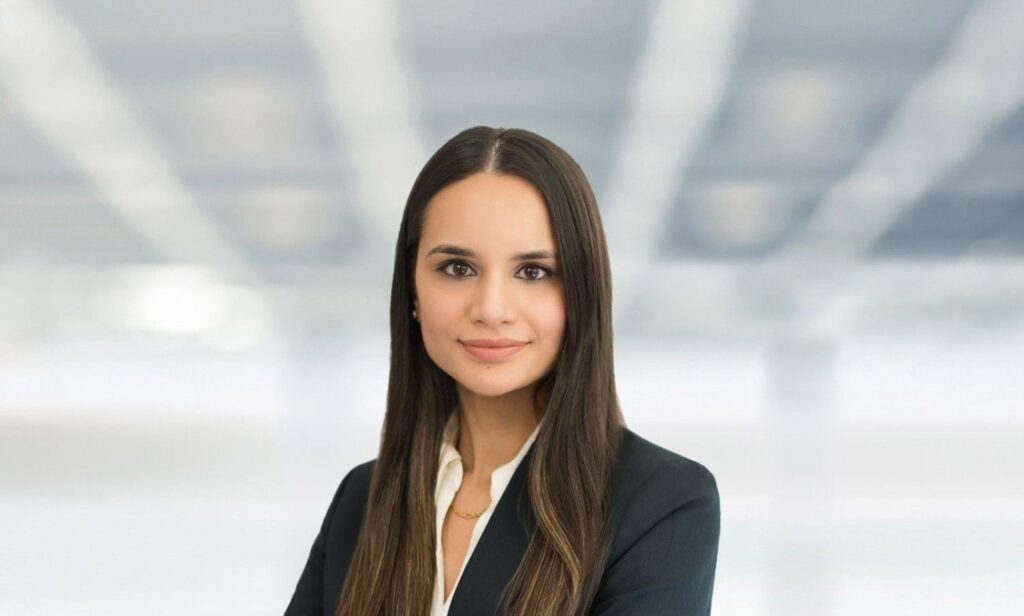 Professional corporate headshot of a young woman in a dark blazer photographed against a blurred architectural ceiling background, taken by Alex Kaplan Photo in Northern New Jersey.