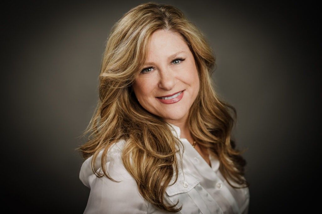 Professional studio headshot of a woman with blonde hair and a natural smile, wearing a white blouse against a neutral gray background, photographed by Alex Kaplan in Northern New Jersey.