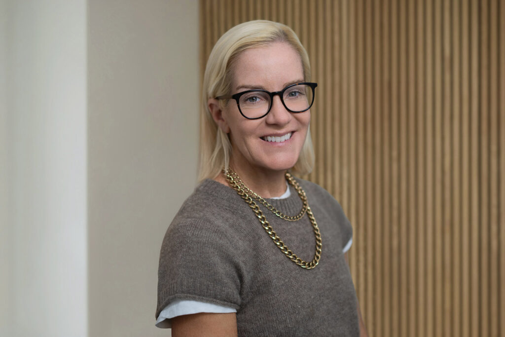 Executive headshot NYC, blonde female executive wearing glasses and gold chain necklace, warm approachable smile, Manhattan office