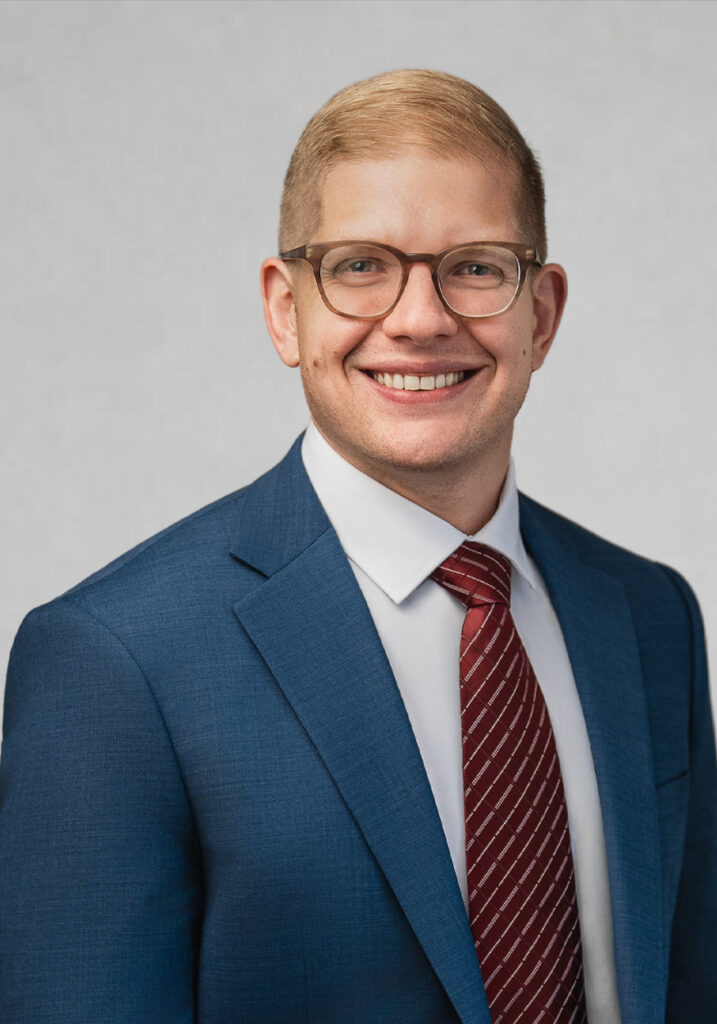 Smiling professional in blue suit, red tie and glasses against a neutral studio background.