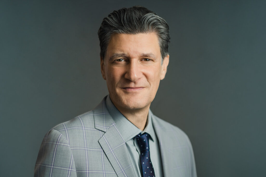Man in a gray suit photographed with directional headshot lighting against a teal background