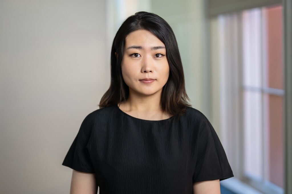 Employee in black textured top photographed for a corporate headshot beside blurred office windows