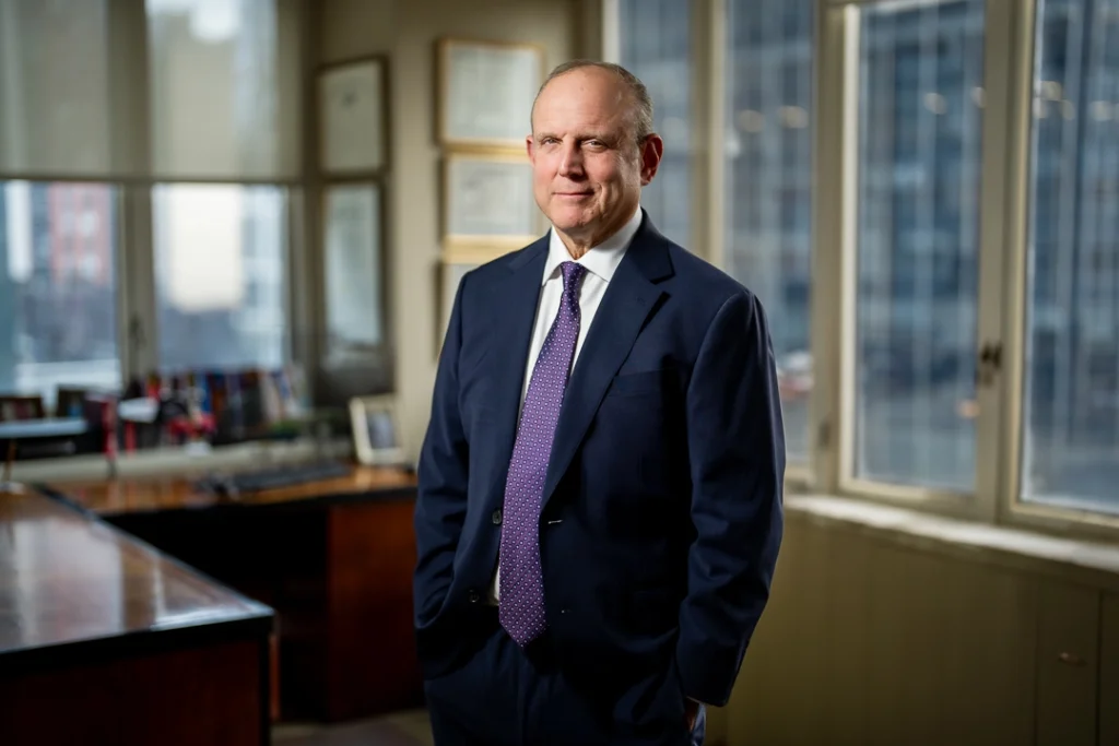 Business professional in a navy suit standing in an office beside windows and a desk